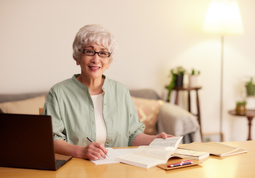 Pessoa idosa estudando em casa com livros abertos e notebook, em um ambiente tranquilo e organizado, favorecendo aprendizado contínuo e bem-estar.